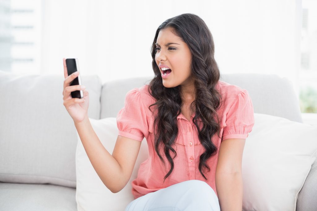 Angry cute brunette sitting on couch holding smartphone in bright living room