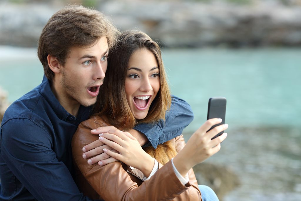 Shocked couple watching a smart phone on holidays on the beach
