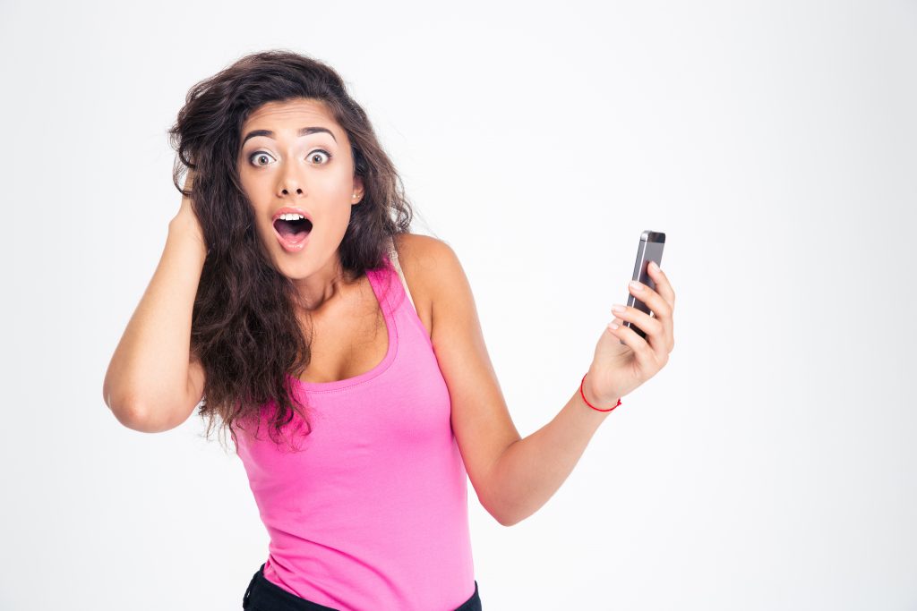 Shocked young woman standing with smartphone and looking at camera isolated on a white background
