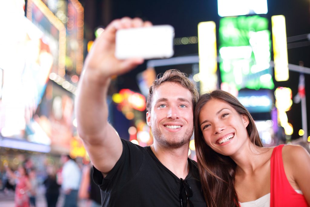 25512692 - dating young couple happy in love taking selfie self-portrait photo on times square, new york city at night. beautiful young tourists having fun date, manhattan, usa. asian woman, caucasian man iPhone 