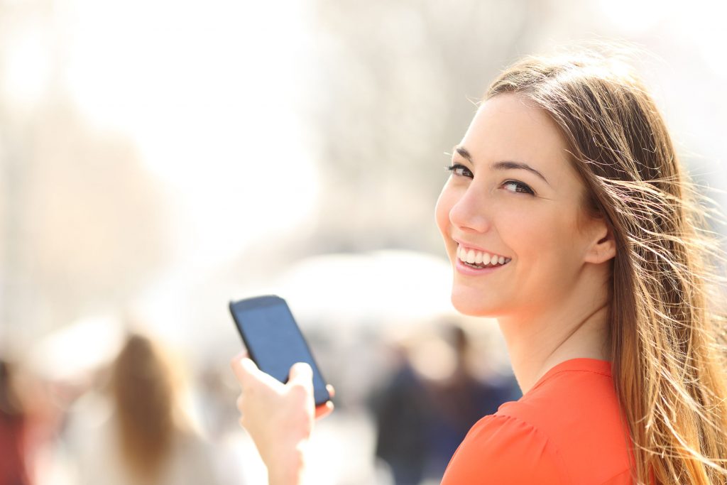 37920383 - happy woman smiling and walking in the street using a smartphone and looking at camera teenager millennials generation y